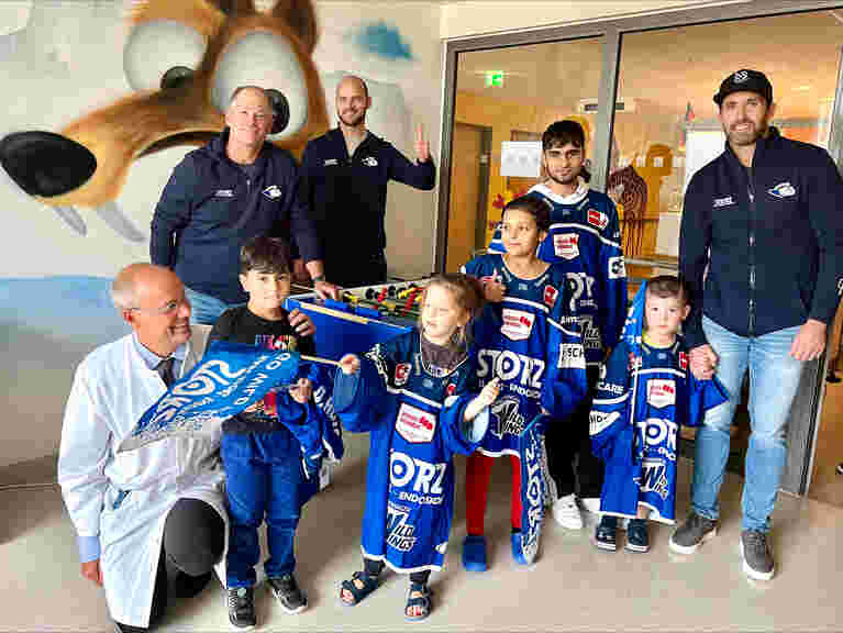 Die vier Wild Wings Spieler in voller Fan-Ausstattung besuchen die Kinderklinik im Schwarzwald-Baar-Klinikum. Sie werden von einem Team von KARL STORZ und einem Arzt begleitet. Die Kinder freuen sich über die Trikots, Fahnen und Pucks.